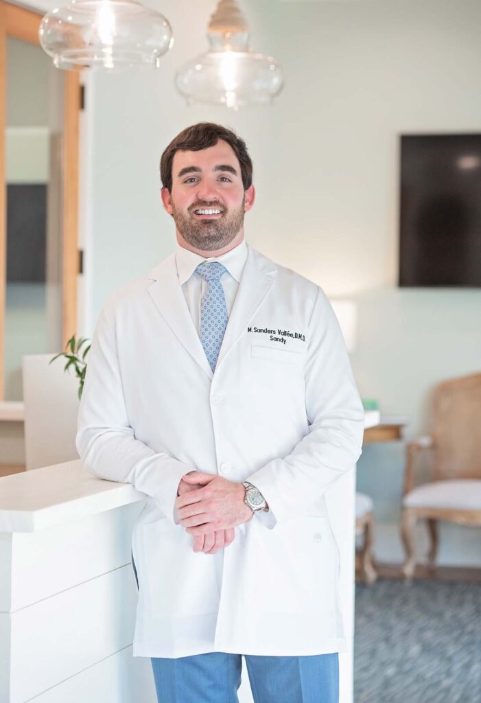 Dr. Sandy Vallee of Eastern Shore Dental Associates in Daphne, AL. They are leaning against a white counter with hands clasped, while the background features a chair, a table with a plant, and a mounted television in a professional office setting.