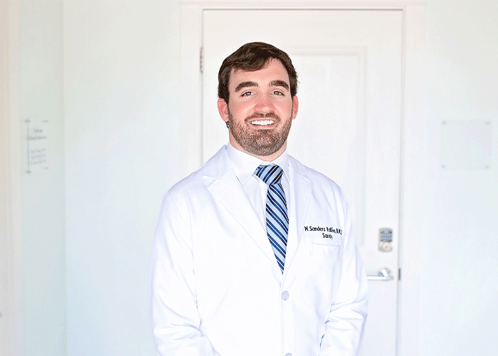Headshot of Dr. Sandy Vallee of Eastern Shore Dental Associates in Daphne, AL. He is wearing a white lab coat with a name tag. They are standing in front of a white door with a keypad lock, dressed professionally with a striped tie.