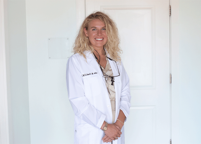 Headshot of Dr. Kalin Roach of Eastern Shore Dental Associates in Daphne, AL. She is wearing a white lab coat, standing in front of a white door. They have curly blonde hair, wear a beige blouse underneath, and accessorize with a cross pendant necklace, glasses hanging around their neck, a watch, and a bracelet, reflecting a professional, likely medical, setting.