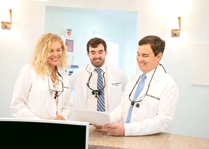 Drs. Roach, Vallee, and Green of Eastern Shore Dental Associates in Daphne, AL. The image features three healthcare professionals in white lab coats gathered around a counter in a medical or dental office. One holds a clipboard, while an AED sign is visible in the background, highlighting a professional and collaborative healthcare setting.
