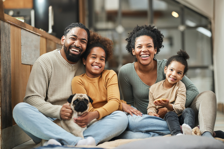 A photo of an African American family sitting together with their dog. Both mom and dad are holding one of their elementary-aged daughters, and the older daughter is holding her dog.