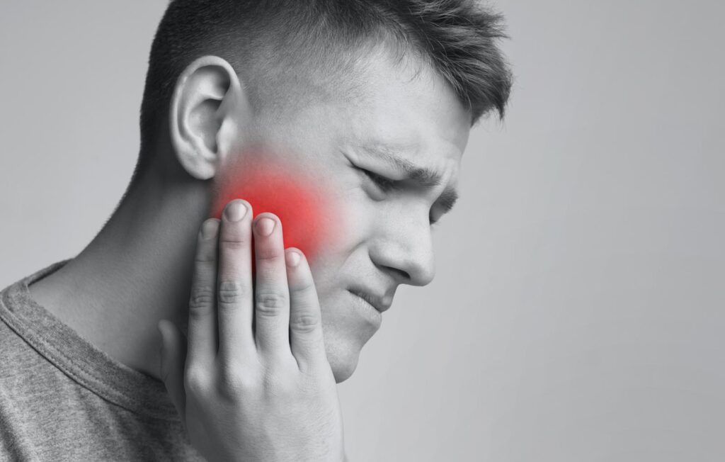 Person holds their cheek with a pained expression, and a red highlight marks the area of discomfort. The image suggests dental or jaw-related inflammation, such as a toothache or TMJ disorder.
