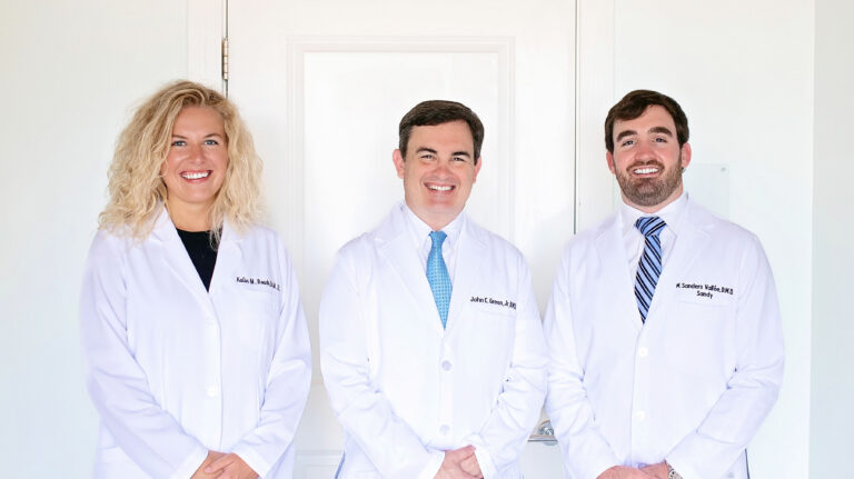 Three dental professionals stand side by side in white lab coats embroidered with their names and titles. They smile in a brightly lit space, suggesting a welcoming and professional team environment.