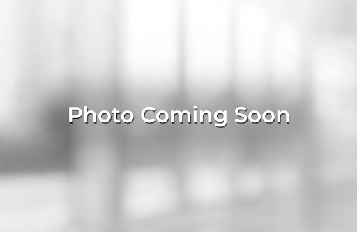 Blurry image shows a single-story building surrounded by trees, with a paved area in the foreground. A sign is visible but unreadable, making the location difficult to identify.